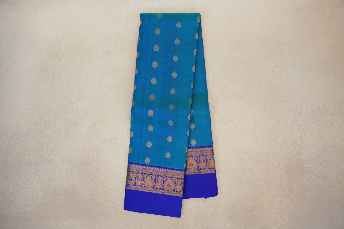 Folded peacock blue lightweight Kanchipuram silk saree with small gold motifs and a royal blue border with gold floral and paisley designs.