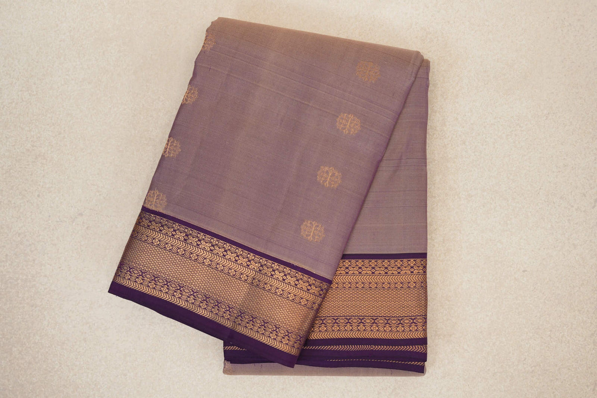 Folded lightweight Kanchipuram silk saree with a muted beige and purple body, scattered gold motifs, and a deep purple border with intricate gold patterns.