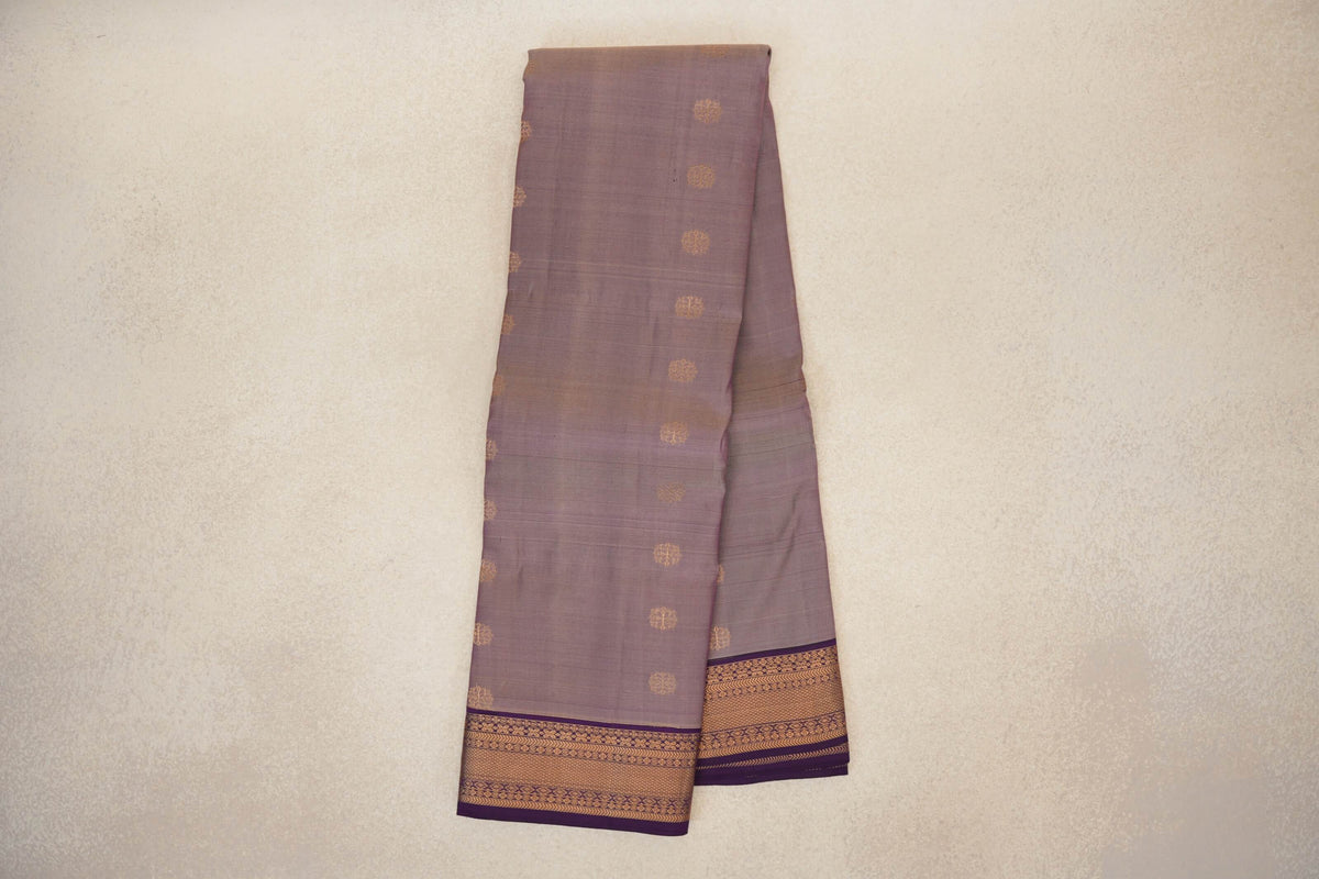 Folded lightweight Kanchipuram silk saree with a muted beige-purple body, small gold motifs and deep purple borders with gold patterning.