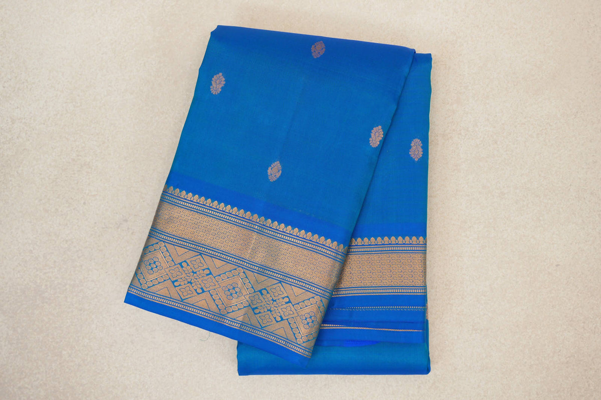 Folded peacock blue lightweight Kanchipuram silk saree with scattered gold motifs and a broad geometric floral border.