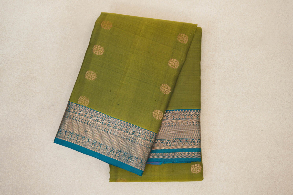 Folded olive green Kanchipuram silk saree with scattered gold-toned round motifs and a teal blue border with intricate woven patterns.