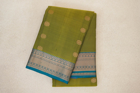 Folded olive green Kanchipuram silk saree with scattered gold-toned round motifs and a teal blue border with intricate woven patterns.