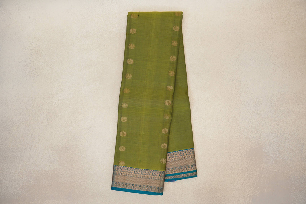 Folded olive green lightweight Kanchipuram silk saree with small gold-toned motifs and a teal blue border with intricate gold-toned patterning.