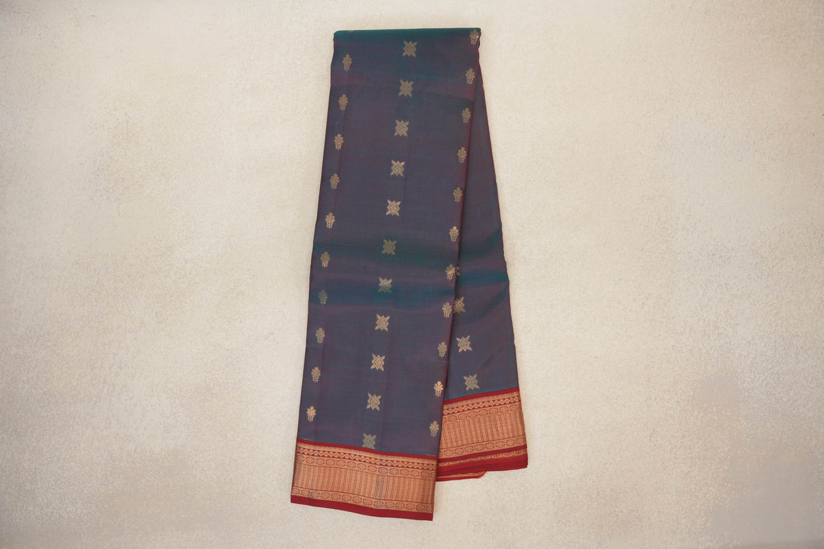 Folded lightweight Kanchipuram silk saree with a dark teal-blue body, scattered gold-toned motifs and deep red patterned borders.