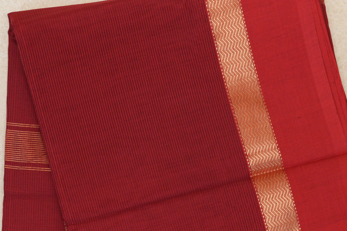 Folded deep red Maheshwari silk cotton saree with fine stripes and a gold-toned zigzag border beside a plain red panel.