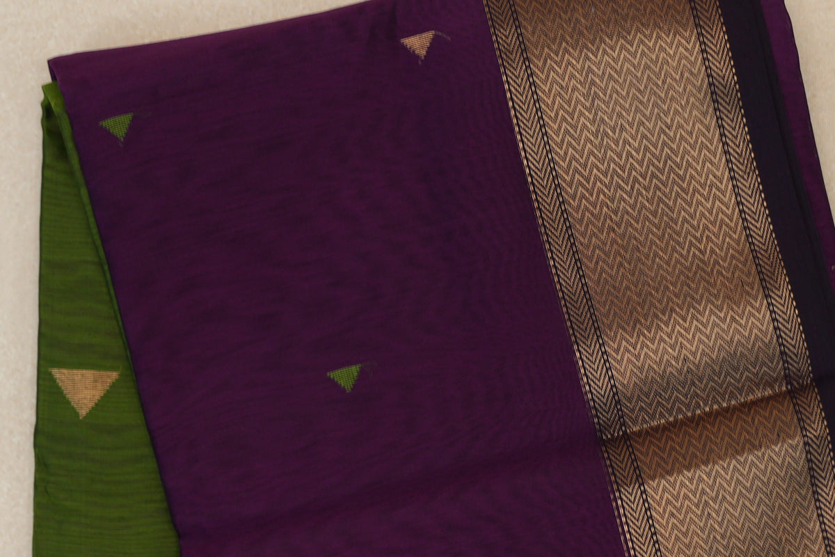 Close-up of a folded deep purple Maheshwari silk cotton saree with small triangular motifs, a broad gold-patterned border and olive green fabric peeking out.