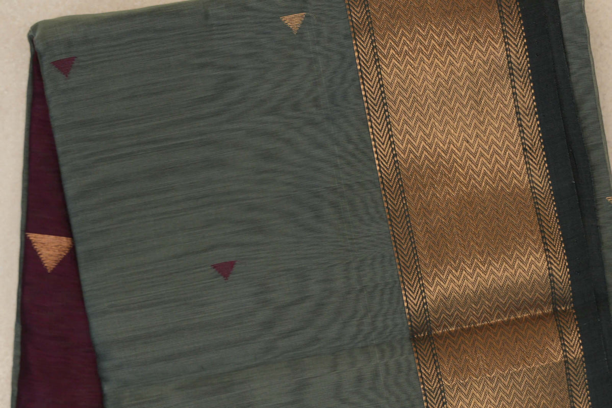 Close-up of a folded sage green Maheshwari silk cotton saree with small triangle motifs, a gold chevron-patterned border and deep wine maroon fabric peeking out.