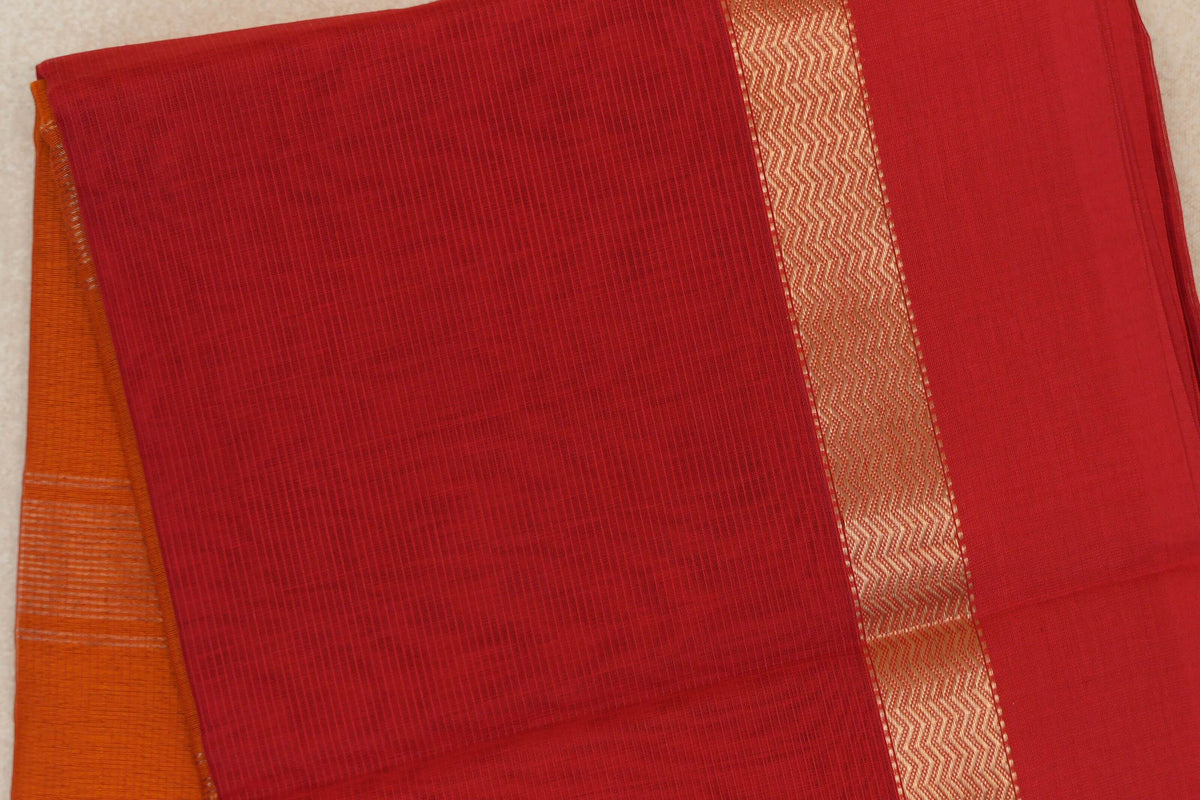 Folded vermilion red Maheshwari silk-cotton saree with fine checks, a gold zigzag border and deep orange fabric peeking out.