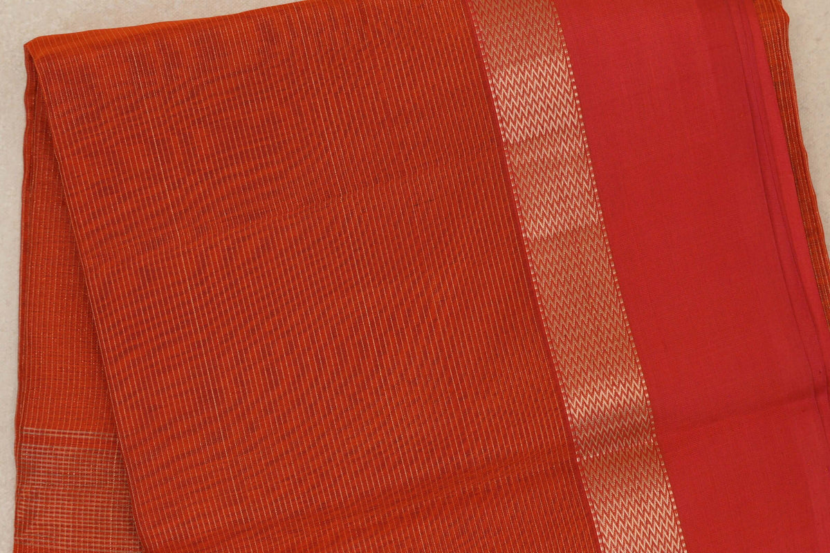 Folded Maheshwari silk cotton saree showing a finely striped rust orange body, a gold-toned zigzag band and a wide red border.