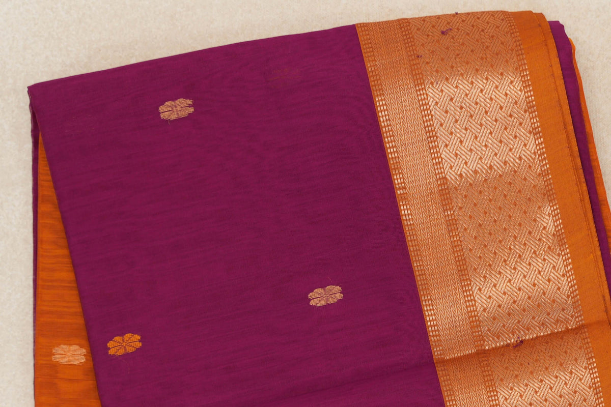 Folded Maheshwari silk cotton saree showing a dark magenta body with small gold motifs and a wide deep orange border with geometric patterns.