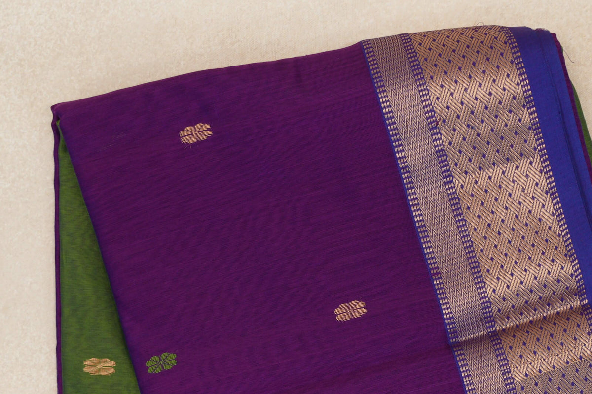 Folded Maheshwari silk cotton saree with a violet purple body, small gold and green motifs, a royal blue border with gold geometric patterns, and olive green fabric beneath.