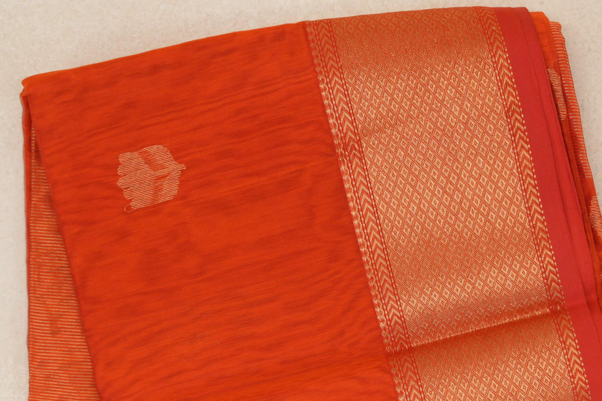 Folded reddish orange Maheshwari silk cotton saree with a plain body, a wide gold-toned geometric border and red selvedge.