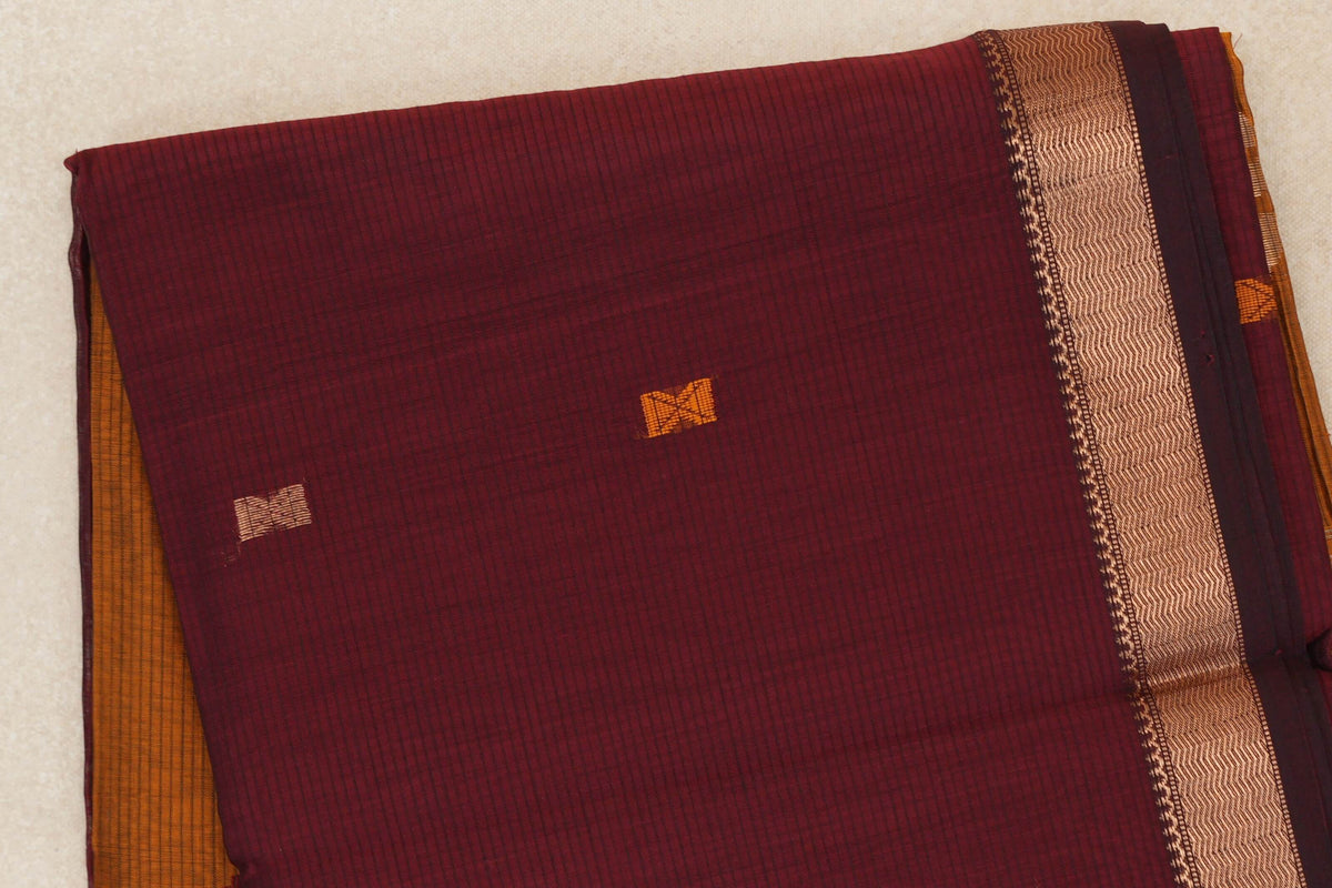 Folded deep maroon Maheshwari silk cotton saree with fine checks, small orange and gold accents, a gold zigzag border and mustard orange fabric peeking out.
