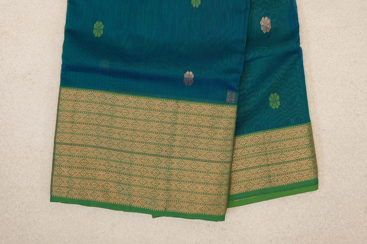 Folded Maheshwari silk cotton saree showing a teal blue body with scattered small motifs and broad leaf green borders with gold-coloured geometric patterns.