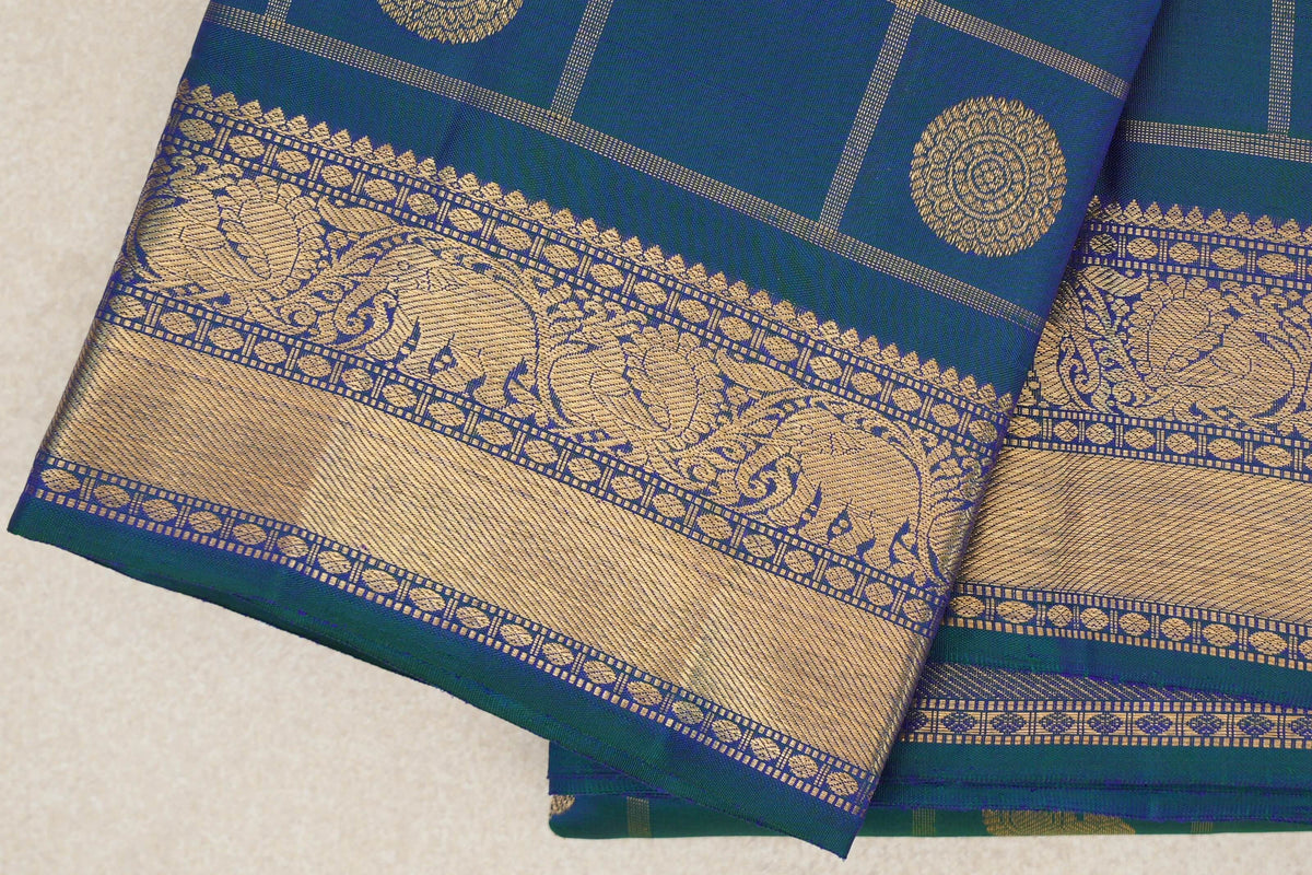 Folded peacock blue Kanjivaram silk saree showing gold circular motifs in a checked body and a broad border with ornate paisley designs.