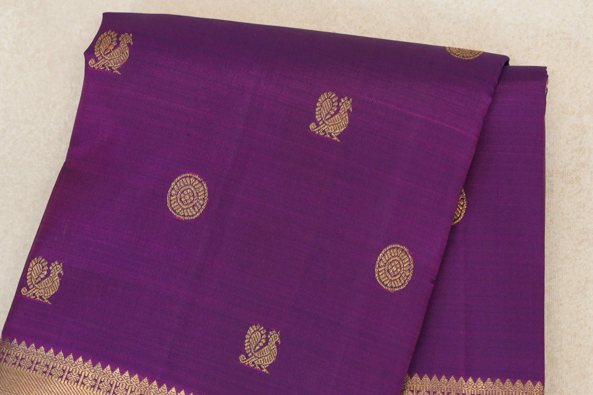 Folded purple Kanjivaram silk saree with gold zari peacock and circular motifs and an ornate border along the lower edge.