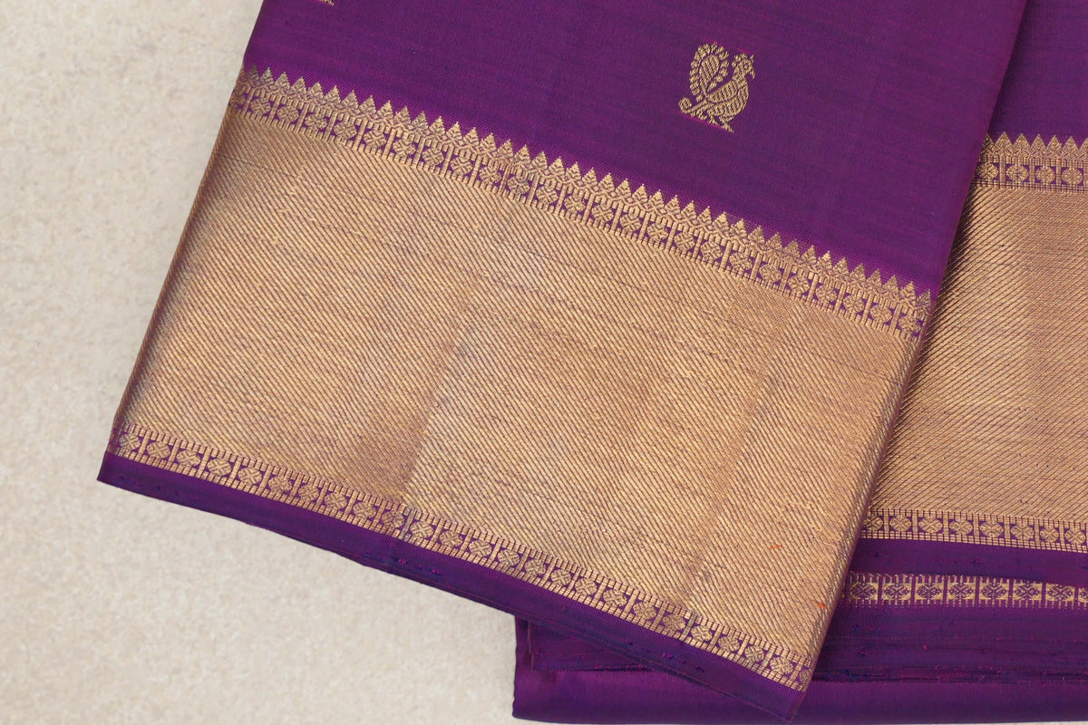Close-up of a folded purple Kanjivaram silk saree with a small gold-toned motif and a wide, finely striped border edged with floral patterns.