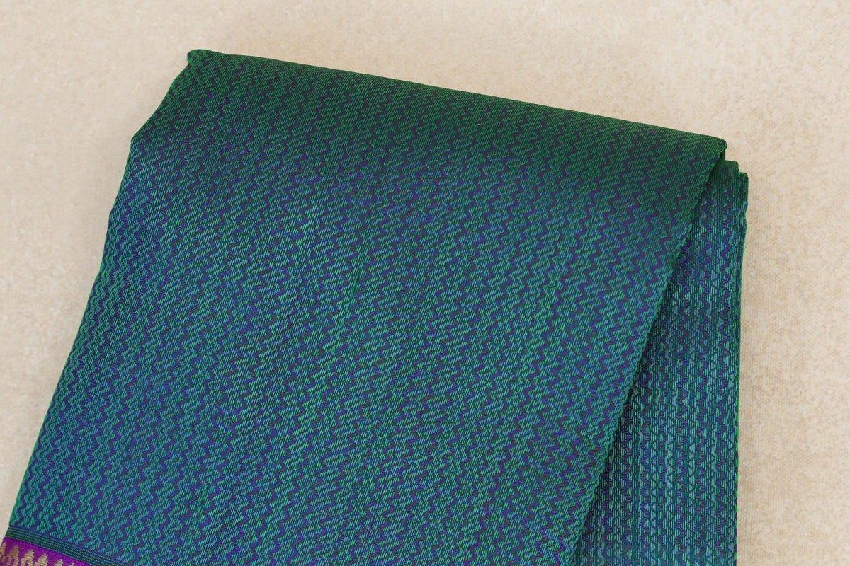 Folded peacock green Kanjivaram silk saree with a fine purple zigzag pattern and a glimpse of its purple border with gold detailing.