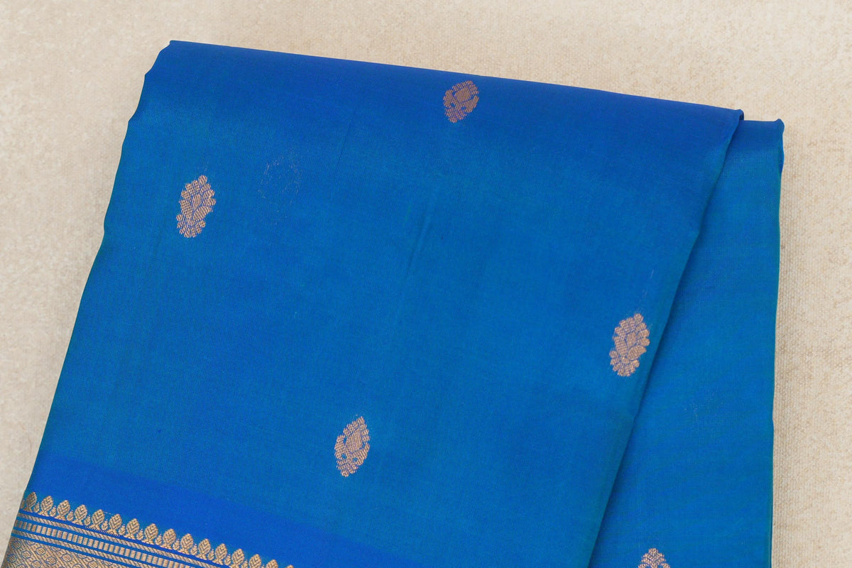 Folded peacock blue lightweight Kanchipuram silk saree with spaced gold zari motifs and a patterned gold border along one edge.