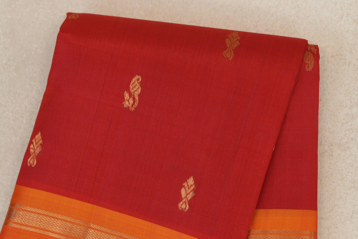 Folded vermilion red light weight Kanchipuram silk saree with scattered gold motifs and a deep orange border with a woven gold pattern.
