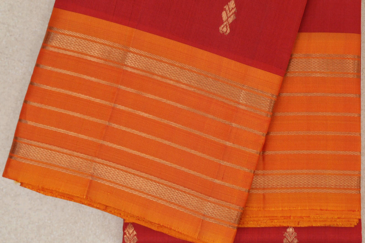 Close-up of a folded vermilion red Kanchipuram silk saree, with small gold motifs and a deep orange border woven with gold bands and fine stripes.