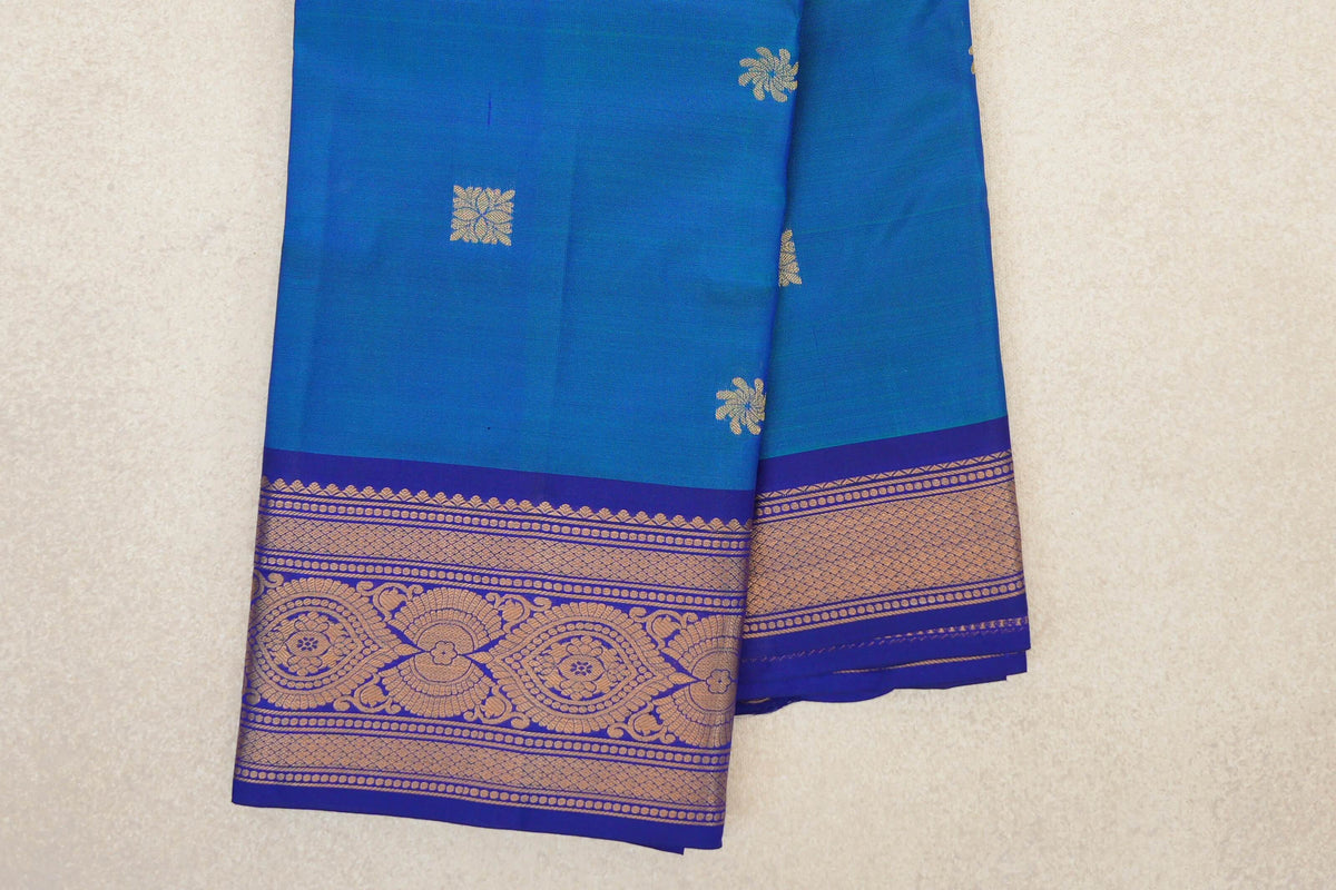 Folded peacock blue lightweight Kanchipuram silk saree with small gold motifs and a deep blue violet border patterned with floral medallions and lattice bands.