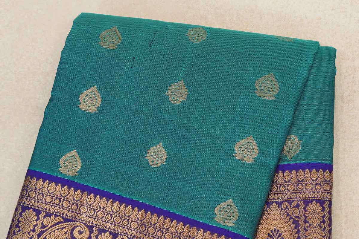 Folded peacock green Kanchipuram silk saree with scattered gold leaf-shaped motifs and a deep royal blue border with gold floral and geometric patterns.