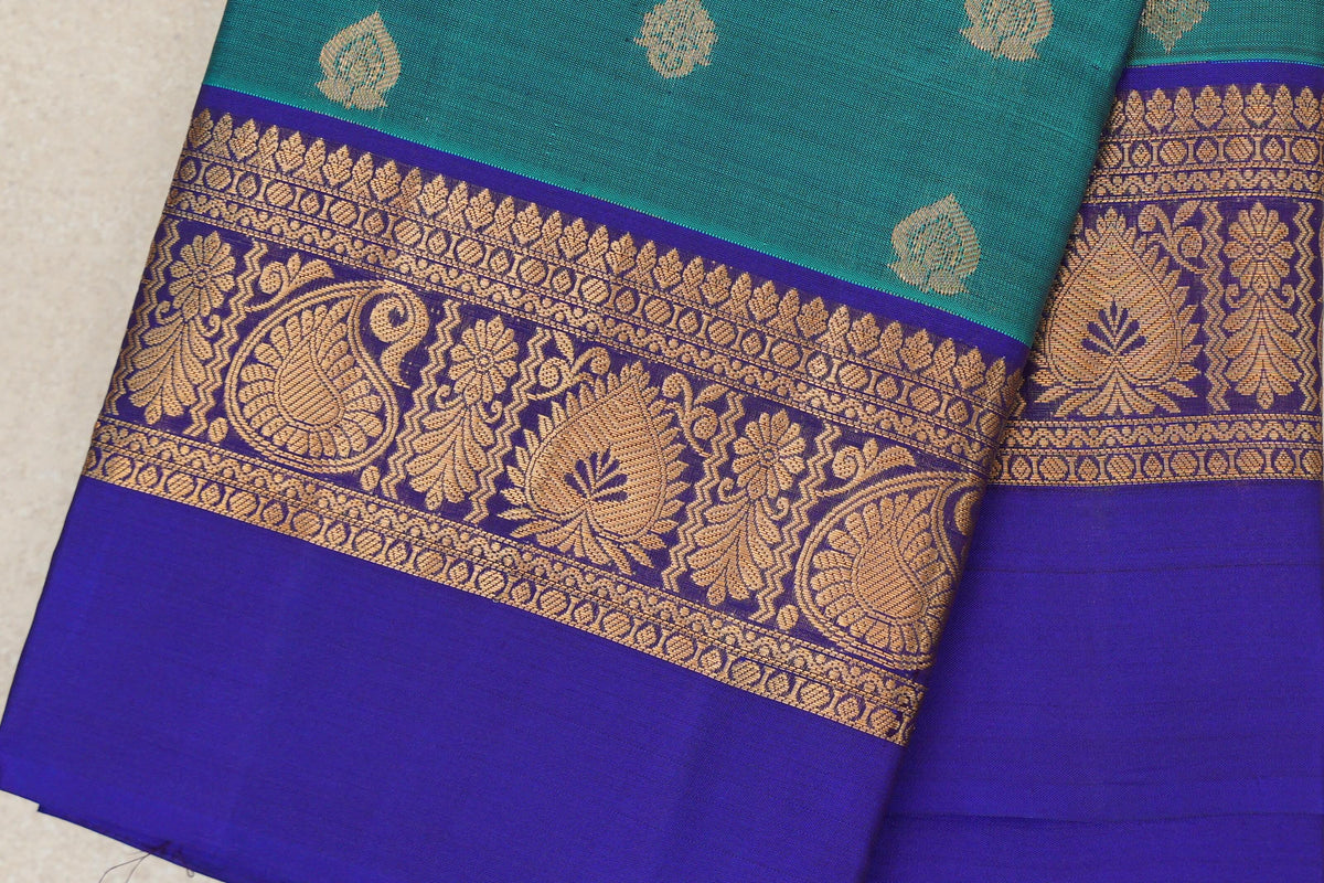 Close-up of a folded peacock green Kanchipuram silk saree with gold leaf motifs and a deep royal blue border woven with gold paisley, bird and floral designs.