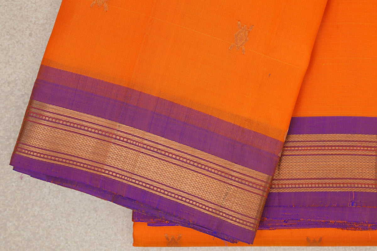 Folded bright orange Kanchipuram silk saree with small gold motifs and a violet border woven with patterned gold bands.