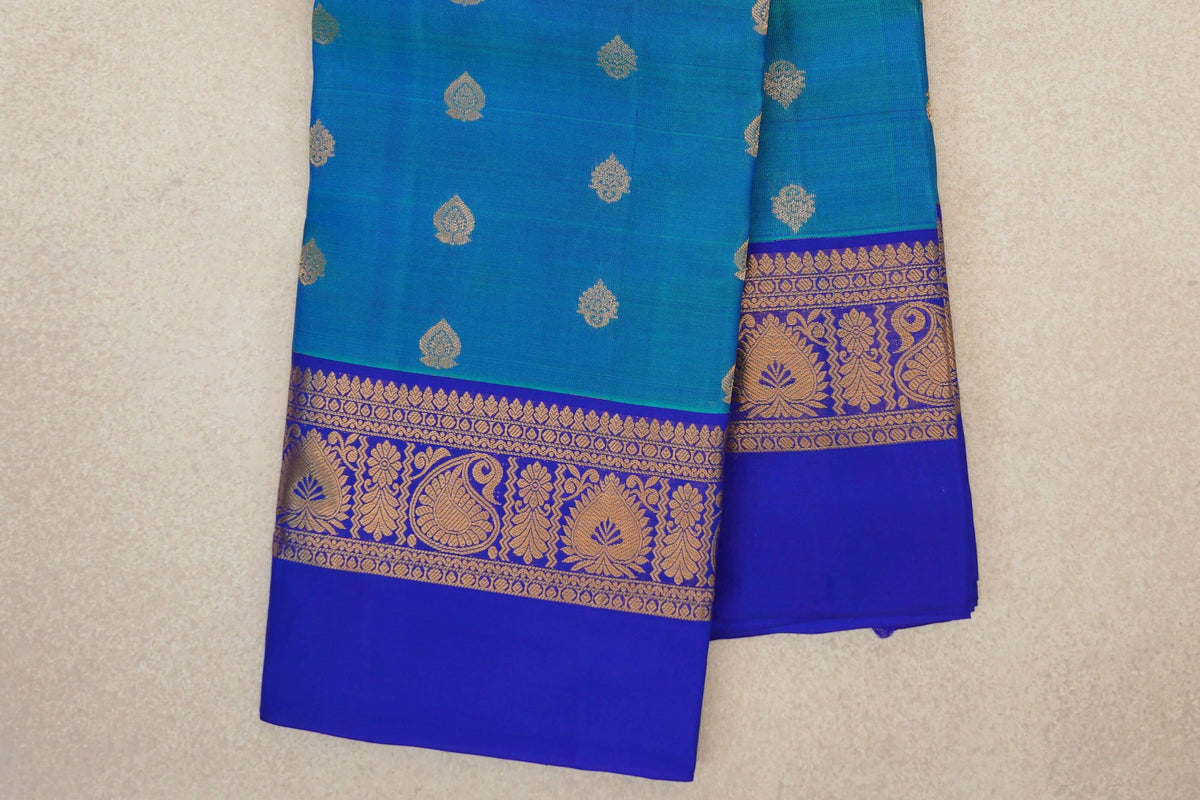 Folded peacock blue Kanchipuram silk saree with scattered gold motifs and a royal blue border featuring gold paisley and floral designs.