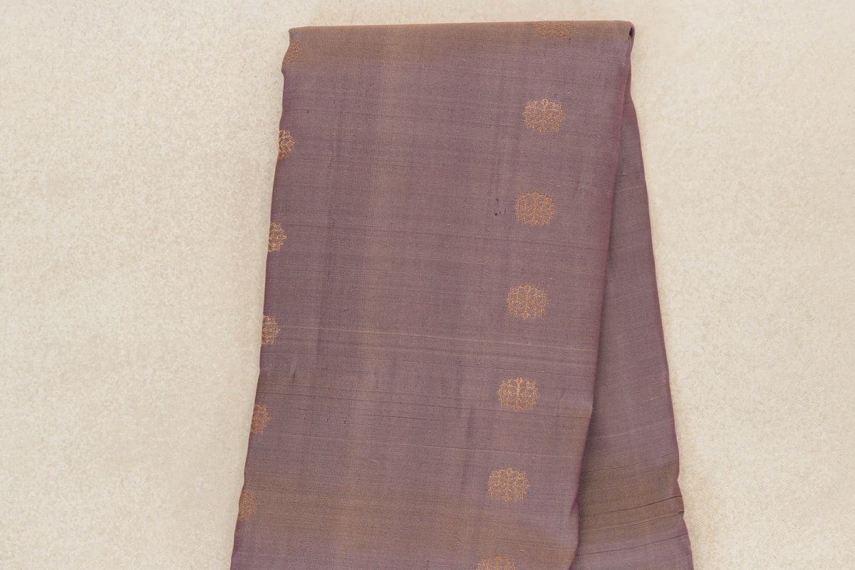 Folded lightweight Kanchipuram silk saree in muted beige and purple, with scattered gold-toned circular motifs and a subtly striped edge.