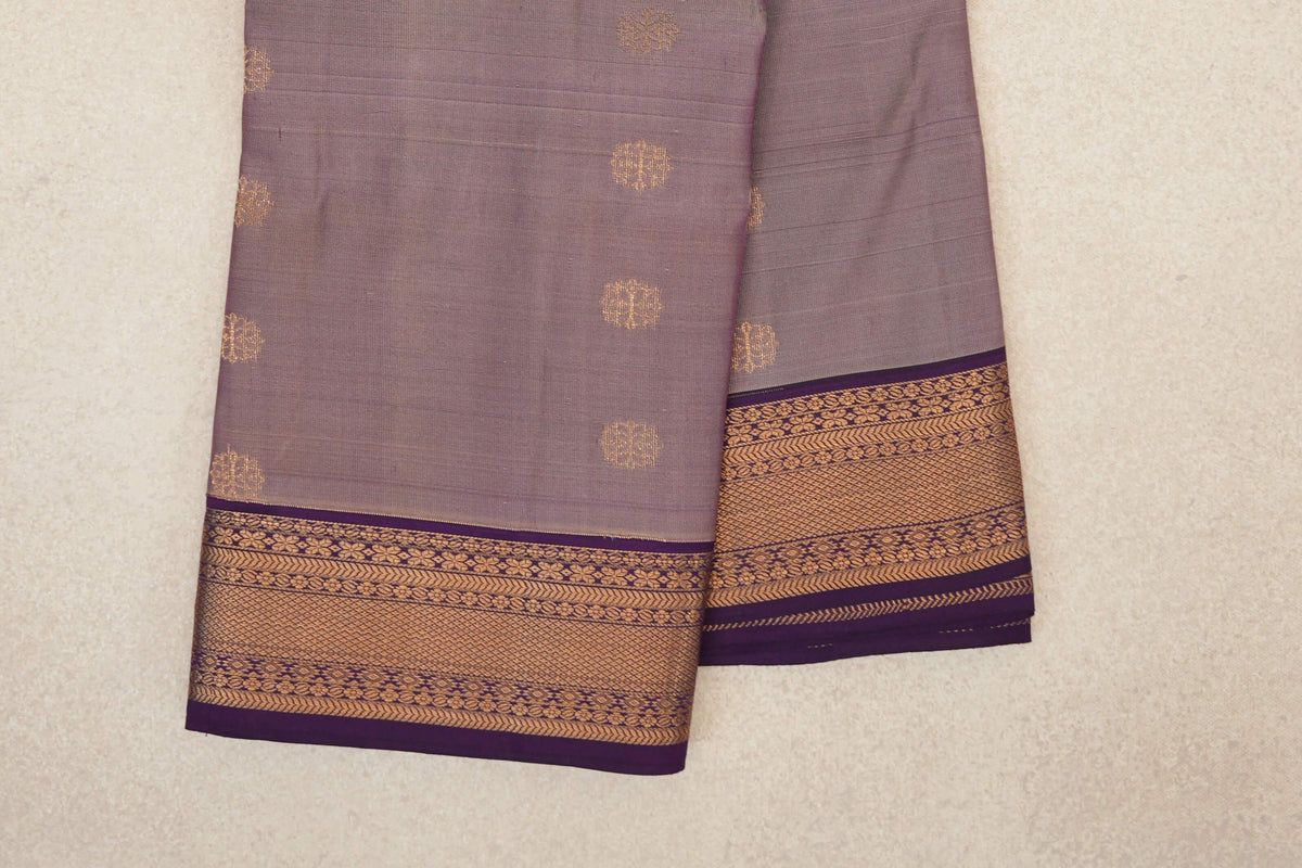 Folded lightweight Kanchipuram silk saree with scattered gold motifs on a muted beige-purple body and ornate deep purple borders.