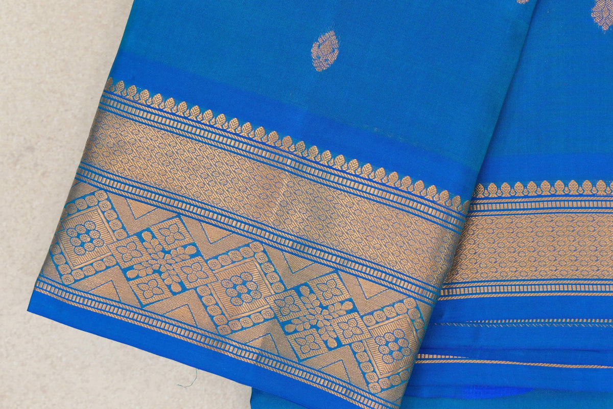 Folded peacock blue Kanchipuram silk saree with small gold motifs and a broad gold zari border of floral and geometric patterns.